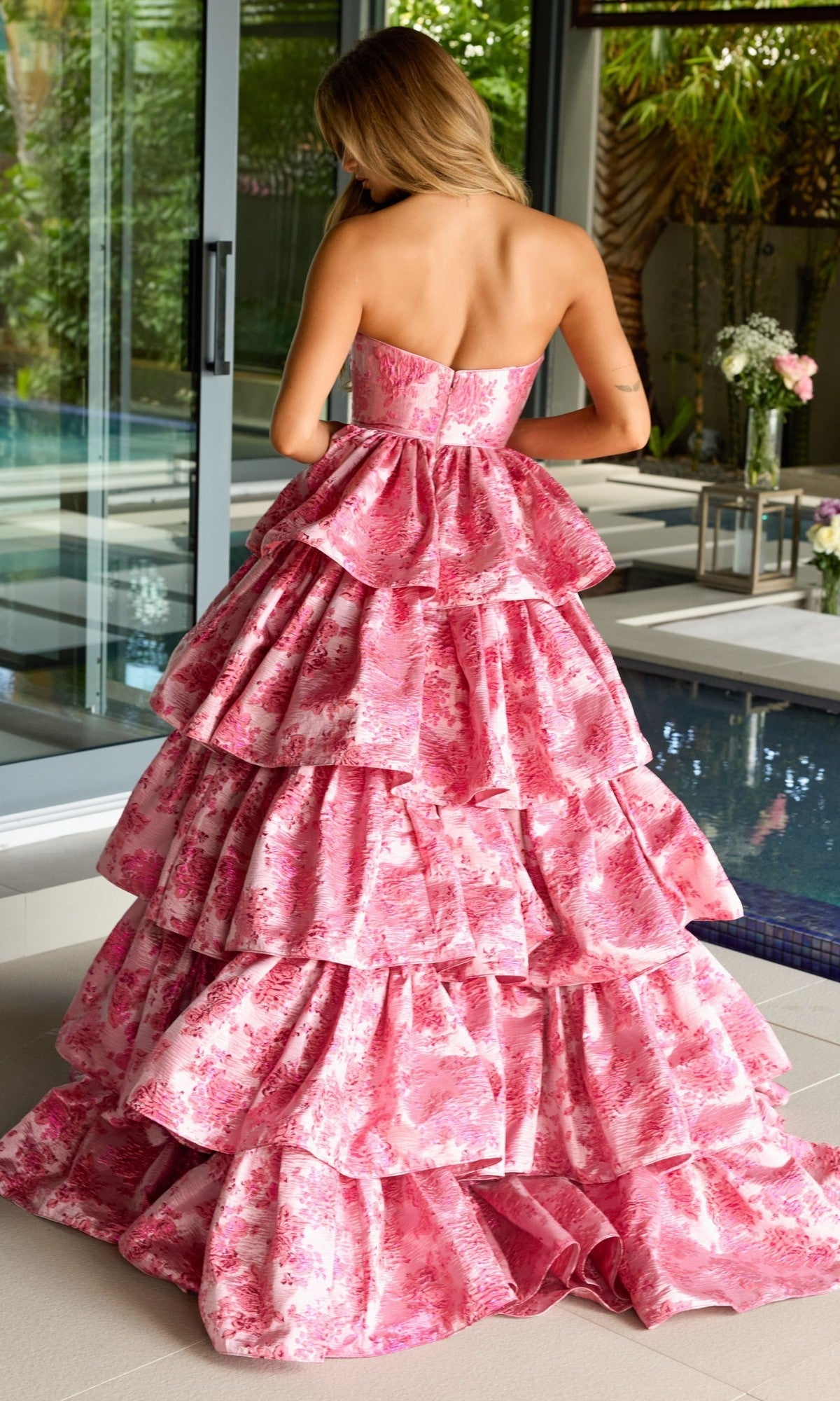 A woman stands indoors by a pool, her back to the camera, wearing the Ava Presley 26412 Ruffled Strapless Prom Gown—a voluminous a-line dress with pink floral print.